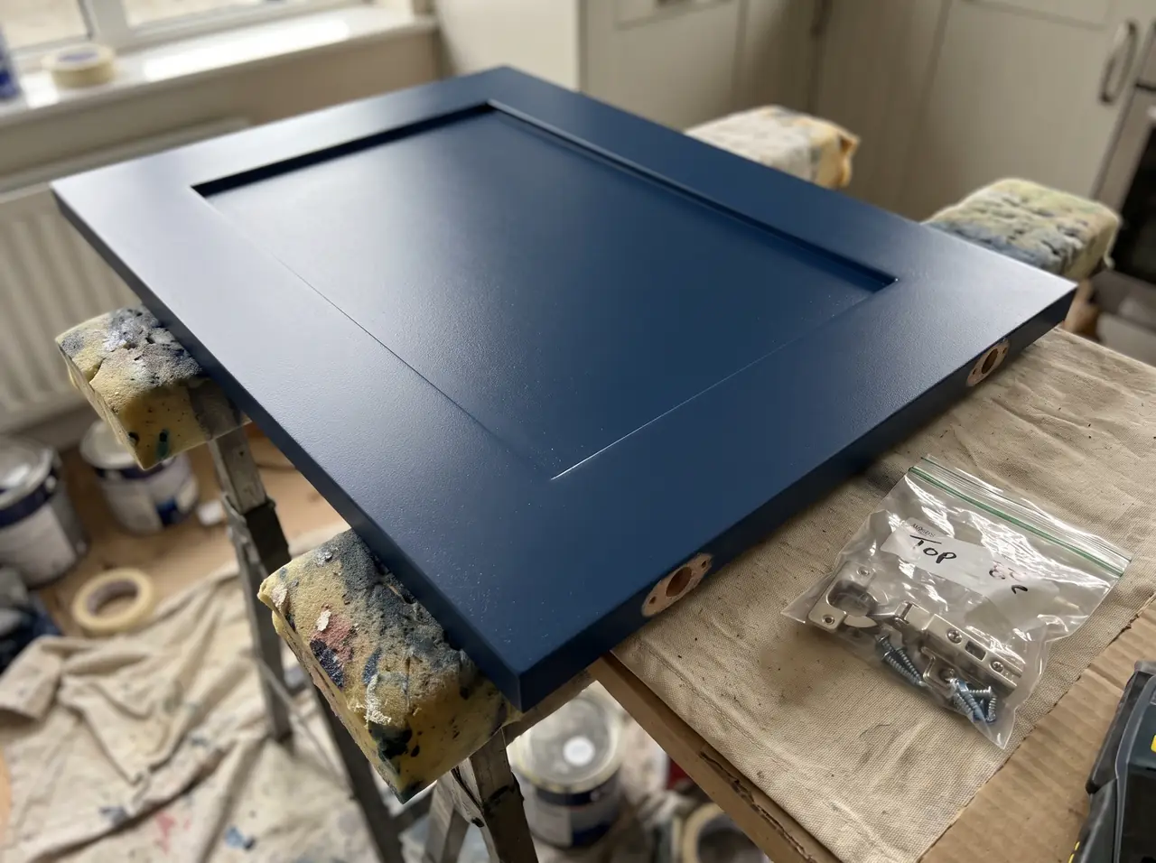 Cabinet door freshly painted navy resting on padded trestles, with its hinges bagged and labelled beside it